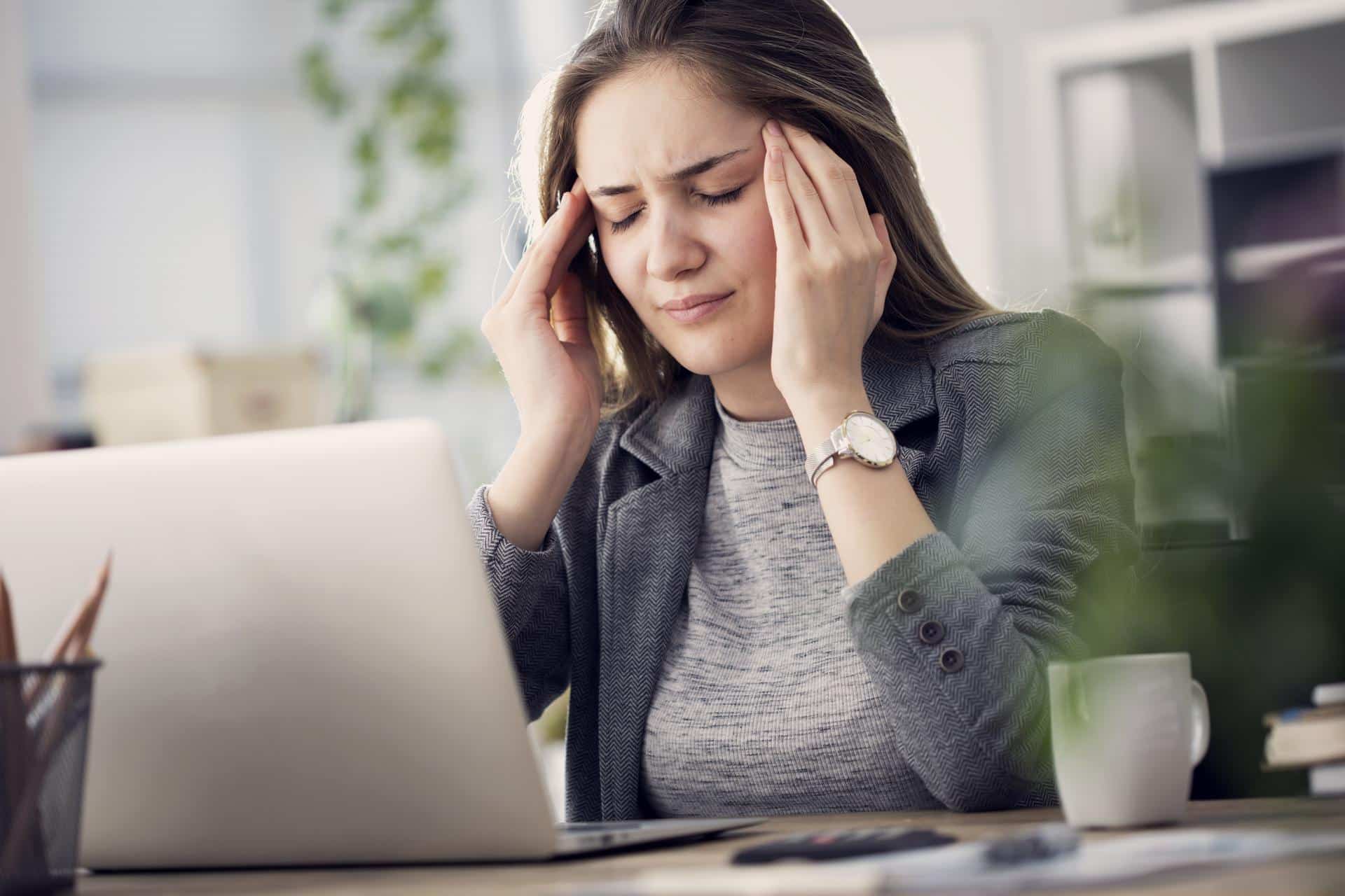 Woman holding her head in pain while working at a computer, showing signs of a tension headache.